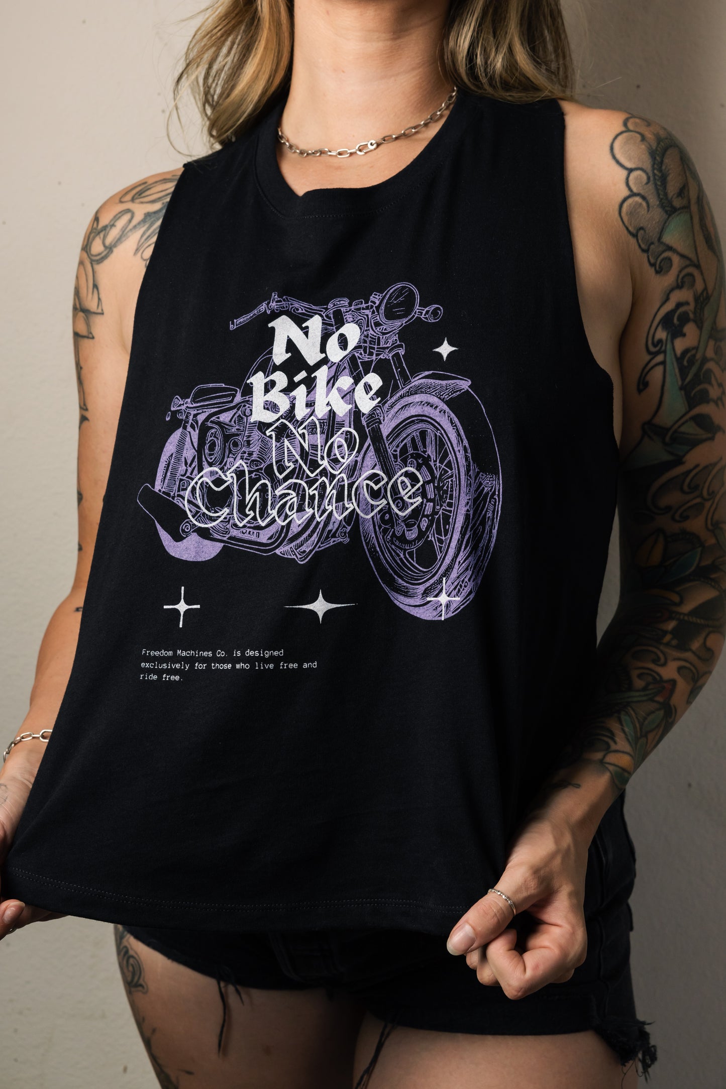 female model with tattoos posing in a black tank top. Tank top has a motorcycle on it with the words no bike no chance. Harley Davidson style streetwear